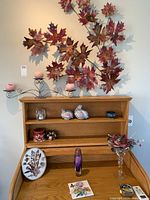 Maple leaf metal wall art with dark red/burgundy leaves above oak roll-top desk holding various decorative items including two metal bird figurines.