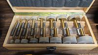 Wooden box containing nine hole saw drill bits with metal shafts and clear variation in sizes.