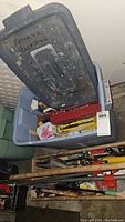 Plastic bin filled with assorted painter's tools, showing container lid with writing and some tools poking out.