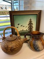 Photo showing the pottery teapot with bamboo handle, multicolored pitcher, and framed tree/bird painting behind.