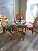 Rattan dining set including round glass top table and three rattan chairs with pink and green cushions in a sunlit room.