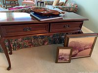 Photo showing dark wood Gibbard sofa table with two front drawers, wooden serving bowl on top, two framed boat scene pictures leaning against table leg, floral sofa background.