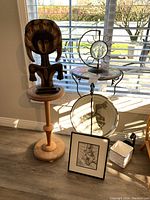 Whole lot viewed together by window. Shows wooden pedestal table with carved wooden figure, metal round table with decorative bowl and glass sun ornament, framed artwork on floor, and small ceramic container.
