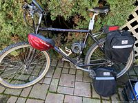 Black Specialized Hard Rock bicycle leaned against foliage on brick ground with accessories including pannier bags attached and helmet on seat.