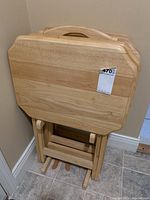 Four wooden folding TV trays nested together on top of their wooden stand on a tile floor against a cream-colored wall. View shows tray tops and part of the stand base.