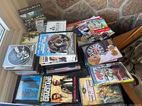 Overall look at the stack of DVDs, CDs, and Nintendo Wii game materials showing most of the lot contents together on a surface against a stone wall background.