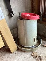Metal cylindrical chicken feeder with red plastic lid and metal tray base, showing wear and some dirt, placed on concrete floor next to wooden board and wall.