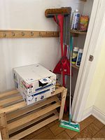 View of wooden slatted shelf with boxes of black garbage bags on top, red broom and dustpan, and a Swiffer mop with green pad in front.