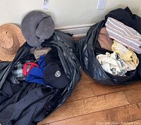 Two large black garbage bags filled with assorted clothing including jackets, shirts, pants, hats, and a grey travel pillow on hardwood floor