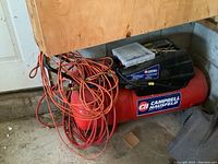 Full view of the red Campbell Hausfeld air compressor on concrete floor in garage, showing orange air hose coiled around unit and black motor on top.
