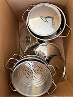 Overview of stacked stainless steel cookware including pots and glass lids inside cardboard box.