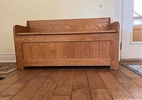 Front view of wooden deacon's bench with panel detailing, showing the bench sitting on a wooden floor.