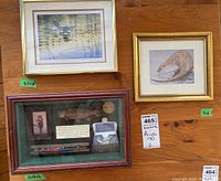 Three items displayed on a wooden table: framed print of loons, framed otter art print, and a wooden shadow box with fishing related items and plaques.