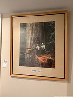 Front view of framed print titled "Foresters," showing two men working on a large tree, with beige matting and light wood frame.