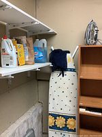 Photo showing ironing board with star and sunflower print cover, Shark brand iron with power cord on wooden shelf, and shelves with detergent bottles