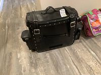 Front view of the black leather motorcycle bag showing buckle straps, studded details, and top handle on wood floor.