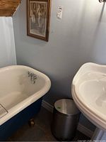 No touch stainless steel trash can positioned beneath a framed print on a wall, next to a vintage bathtub and pedestal sink in a bathroom corner.