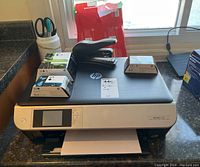 Front view of HP Envy 5530 printer with three packs of HP 61 cartridges on top, black stapler, scissors, and paper loaded in printer tray.