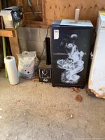 Full view of the black Bradley smoker unit in a garage setting with a vent pipe on the side, thermometer on door, and control panel visible.