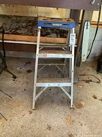 Front view of Featherlite aluminum step ladder showing two steps, blue top, and metal frame standing on a concrete floor with surrounding garage items.
