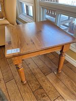 Square wooden side table placed near a window, showing wood grain and light scratches on the tabletop.