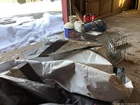 Photo showing a large black and gray tarp folded on the ground with a rectangular metal live trap cage beside it inside a garage area.