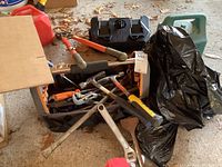 Photo showing a black plastic toolbox filled with assorted hand tools including wrenches, hammers, and bolt cutters with orange handles. Joined by some black bags and a green Coleman lantern beside.