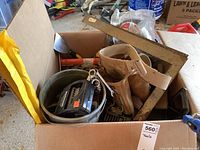 Overview of cardboard box containing assorted tools including tool belt, T-square ruler, and a metal bucket containing various hand tools.