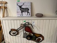 Photo shows metal motorcycle wall art mounted on white wall above a cabinet, next to the moose canvas print and a natural rock.