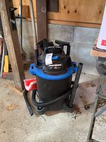 Mastervac wet/dry vacuum in black and blue, standing on a concrete floor with attached hose and some tools beside it.