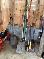 Multiple yard tools including two shovels, a broom, an axe, and a hoe leaning against a wooden wall inside a garage.