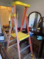 Photo of a 9 ft orange and yellow aluminum step ladder with a yellow top tool tray placed indoors next to furniture.