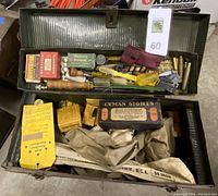 Open toolbox displaying gun cleaning supplies, boxes, wooden handles, brass cartridges, tools, and packaging inside two-tier compartments.