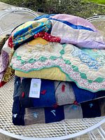 Stack of four folded quilts showing various patterns and textures including floral hexagons and patchwork squares.