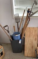 Photo of a large lot of lawn tools including pitchforks, shovels, hoes, small cultivator, and two plastic containers in a gray bin showing condition and arrangement of tools.