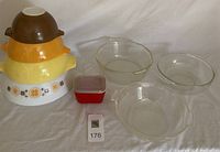 Photo showing a stack of nested Pyrex mixing bowls in brown, orange, yellow, and patterned white, a small red container, three clear glass mixing bowls, and a clear glass refrigerator dish.