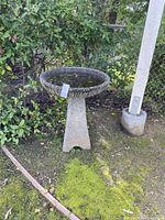 Concrete mid-century modern style birdbath standing on grass near a fence, showing its round basin and pedestal base with no visible damage.