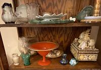 Shelf with various glassware, ceramic, and decorative items including shell collection in glass container, orange pedestal dish, and pearl-shell trinket box