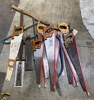 Top down view of a pile of saws including 9 traditional wooden handle hand saws and 4 hacksaws with metal frames and handles