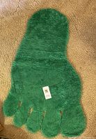 Green shag rug shaped like a large foot with defined toes, laid flat on a carpeted floor showing plush texture and bright green color.