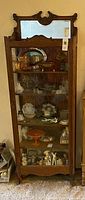 Full front view of vintage oak curio cabinet filled with items, showing ornate carved wood trim at top with mirror and glass shelves.