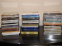 Three stacks of assorted music CDs by various artists on a dark surface against a plain background.