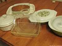 Two clear Pyrex glass dishes are displayed front and center including one round pie plate with a flower sunflower pattern bottom and one rectangular loaf pan. Behind are two green Pyrex dishes including a Daisy pattern baking dish with lid. Also present are Corningware French White round and rectangular casserole dishes along with a white ovenware dish with lid and a woven bun basket.