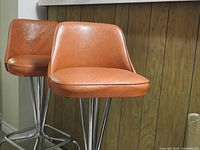 Two vintage orange pleather swivel bar stools with chrome bases positioned side by side against wood panel wall.