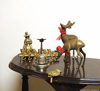 Wide angle image showing entire lot on wooden table including pair of etched brass reindeer, brass cat coat hook, Welsh Pilgrim Lady figurine, etched brass bell, and pineapple key holder.