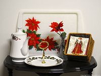 Photo showing vintage porcelain teapot with holly design, large rectangular poinsettia serving tray, bone china sweets tray with golden handle, wicker basket with Christmas napkins featuring Santa Claus, and pressed glass vase with silk poinsettia flower all arranged on a black table.
