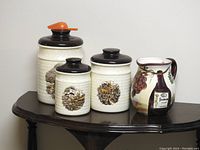Full view of three beige ceramic canisters with red scoop and a Casa Elite pottery pitcher on wooden table.