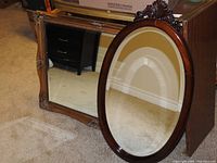 Large rectangular gold-framed mirror leaned on carpeted floor alongside a dark wood oval mirror.