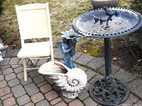 Cast iron birdbath with central bird-on-branch ornament, ornate pedestal, concrete seashell planter with peeling paint, fairy plant stand, and children's chair outdoors on brick and stone patio.