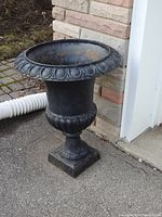 Side view of one black metal garden urn showing decorative rim and scalloped base with rusting inside.